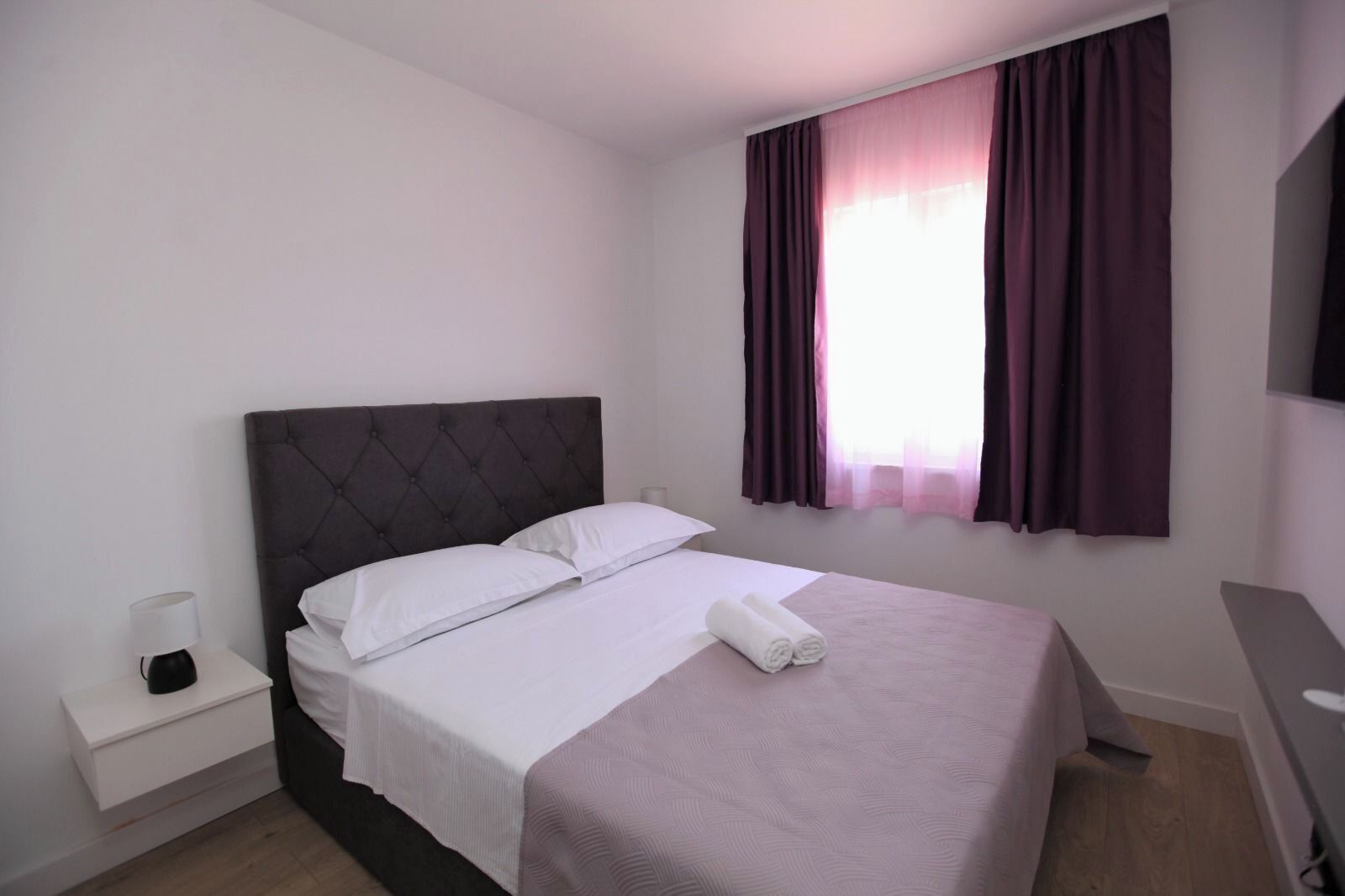 A neatly made bed with white linens and a dark headboard, a bedside table with a lamp, and a window with purple curtains.