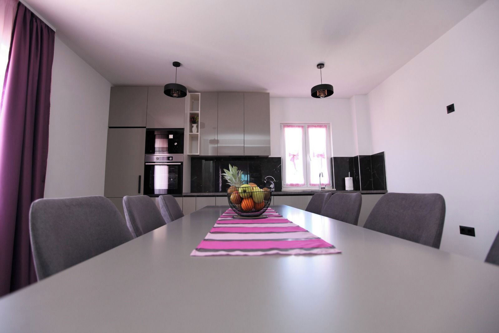 Un espace de salle à manger moderne avec une longue table, des chaises grises, un centre de table composé d’un saladier de fruits, et une cuisine en arrière-plan.