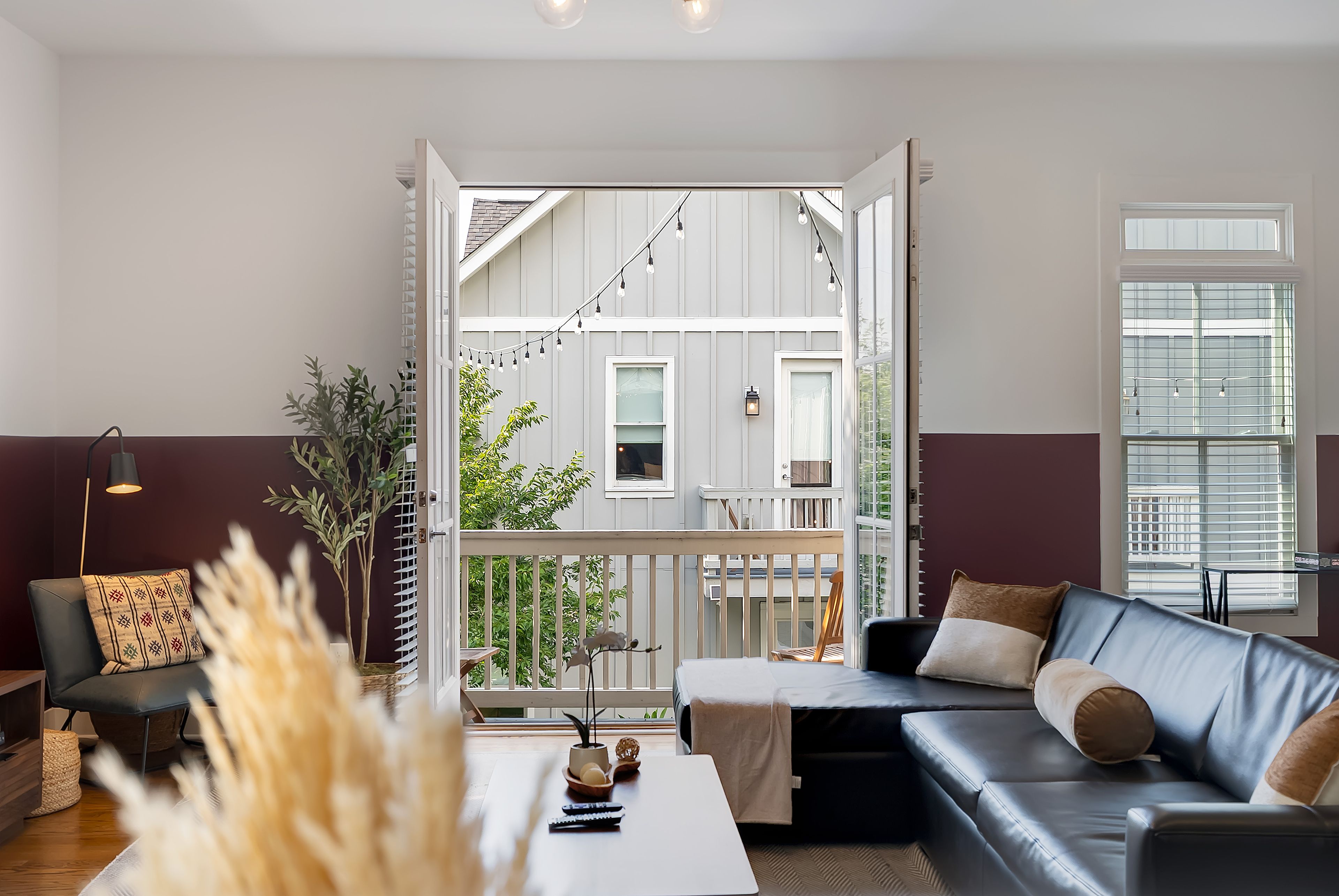 A modern living room with a black leather sofa, a wooden coffee table, and a view of a balcony with string lights.