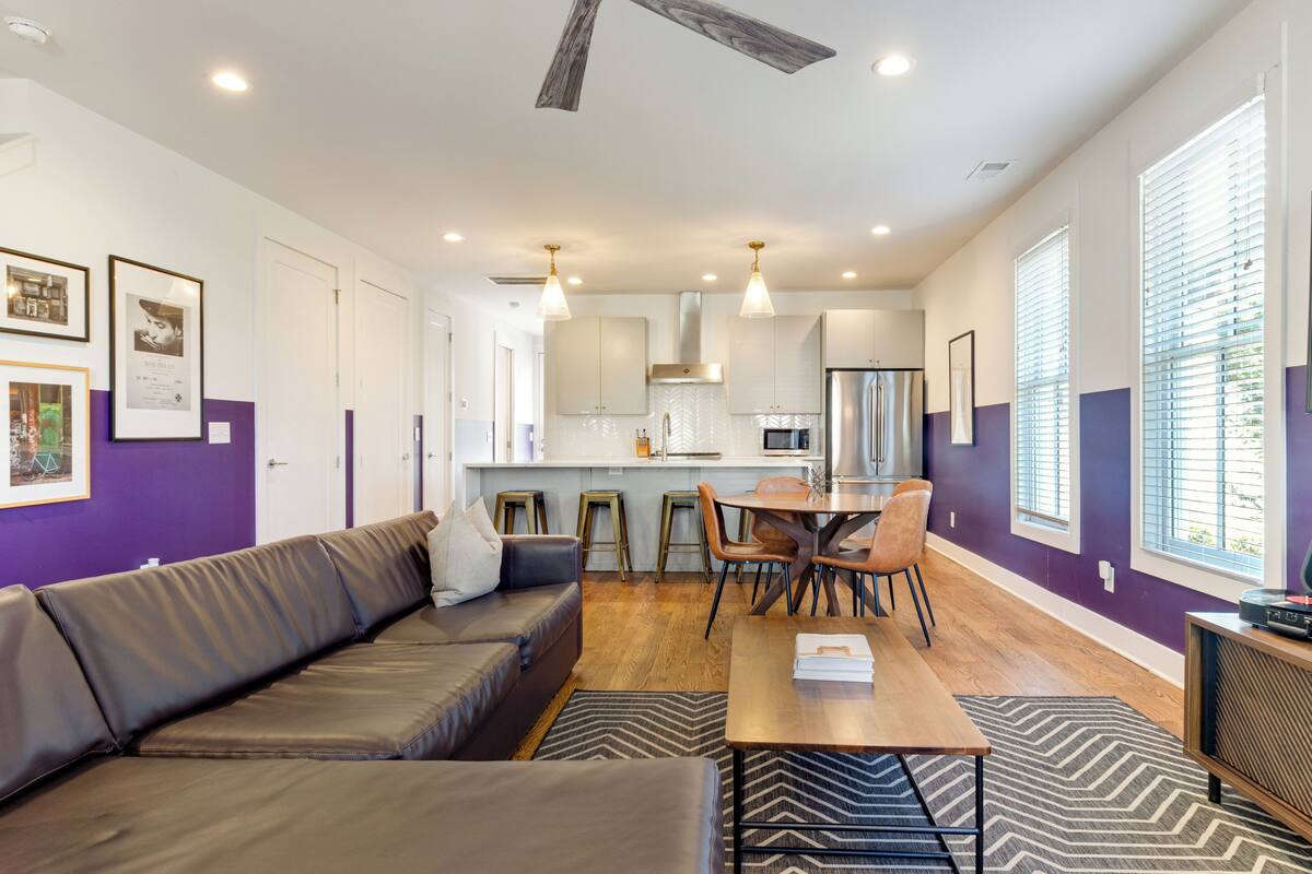 A modern living room with a sofa, dining table, and chairs. There is a kitchen area with a refrigerator, microwave, and stove. The room has large windows with blinds and a ceiling fan.