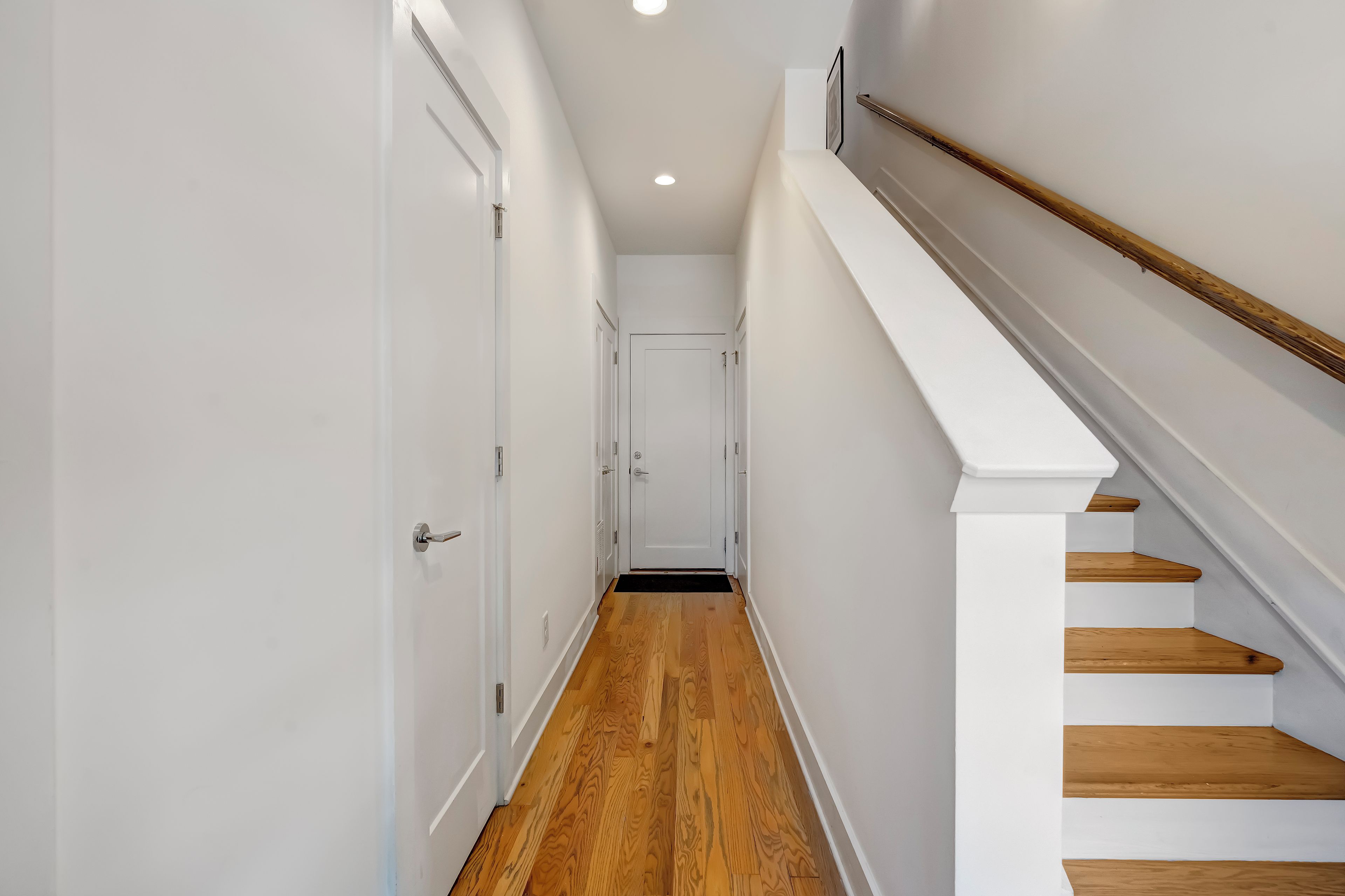 Un pasillo estrecho con piso de madera, paredes blancas y una escalera blanca con escalones de madera.