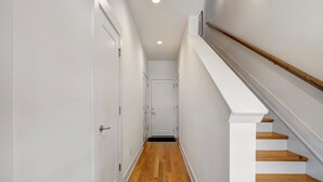 Un pasillo estrecho con piso de madera, paredes blancas y una escalera blanca con escalones de madera.