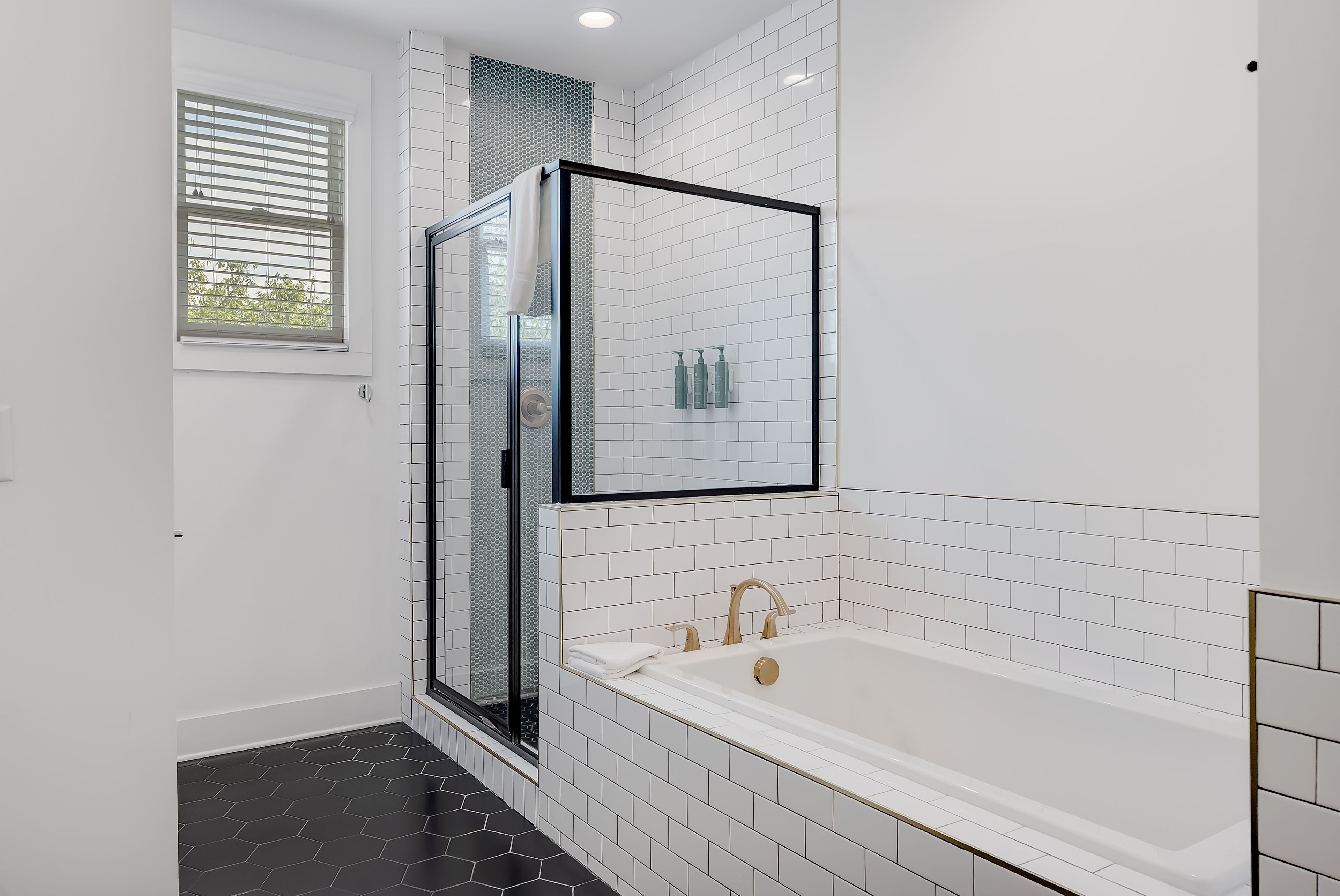 A modern bathroom with a glass-enclosed shower, a white bathtub, and a window with blinds.