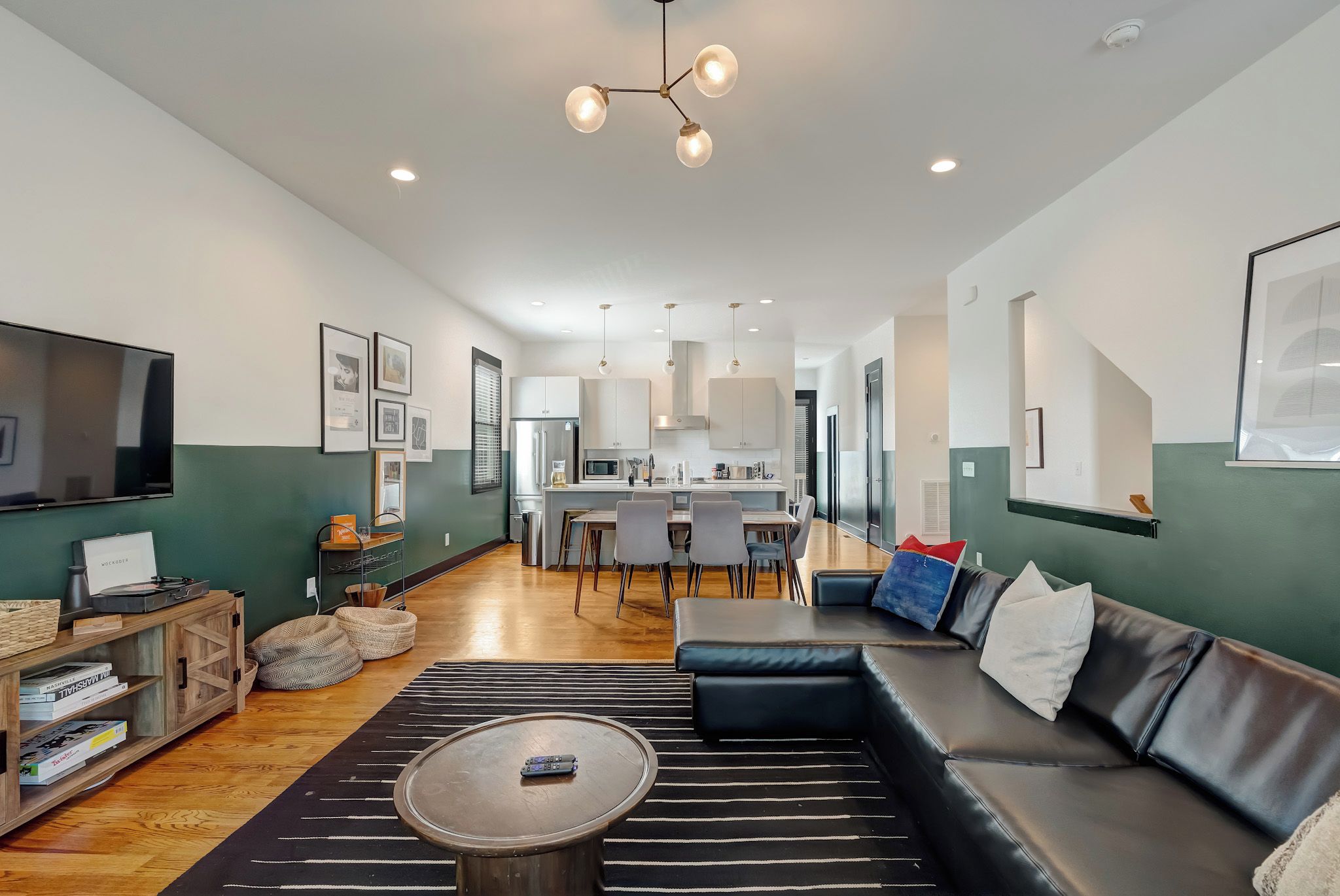 A modern living room with a sofa, coffee table, and a TV. There is a kitchen in the background with a dining area.