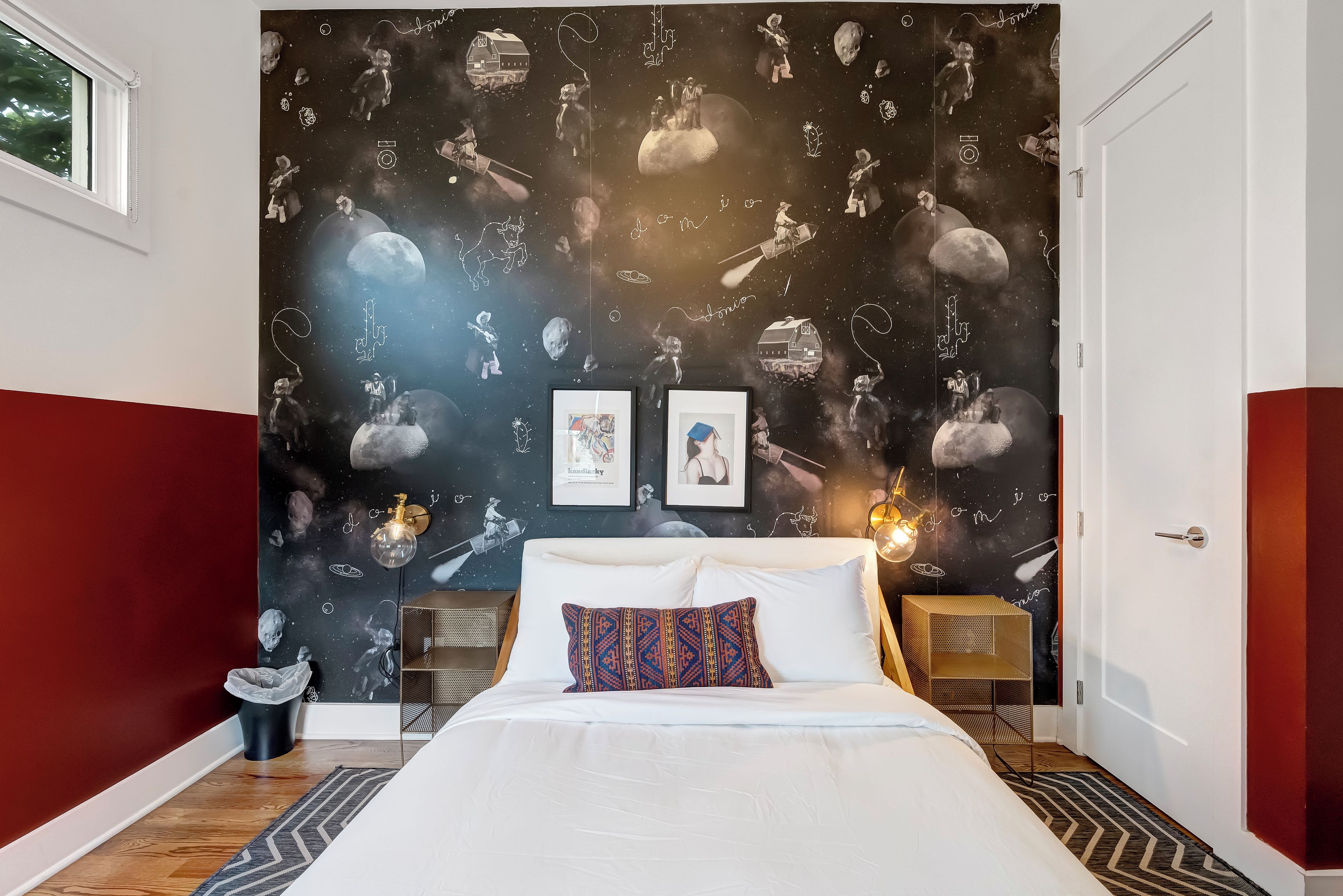Un dormitorio con una cama, dos mesitas de noche, una mesita auxiliar, una pared con un mural de temática espacial y una ventana.