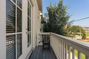 A wooden deck with a white railing, a black chair, and a clear view of a tree and a building in the background. - Monroe Houses (Nashville)