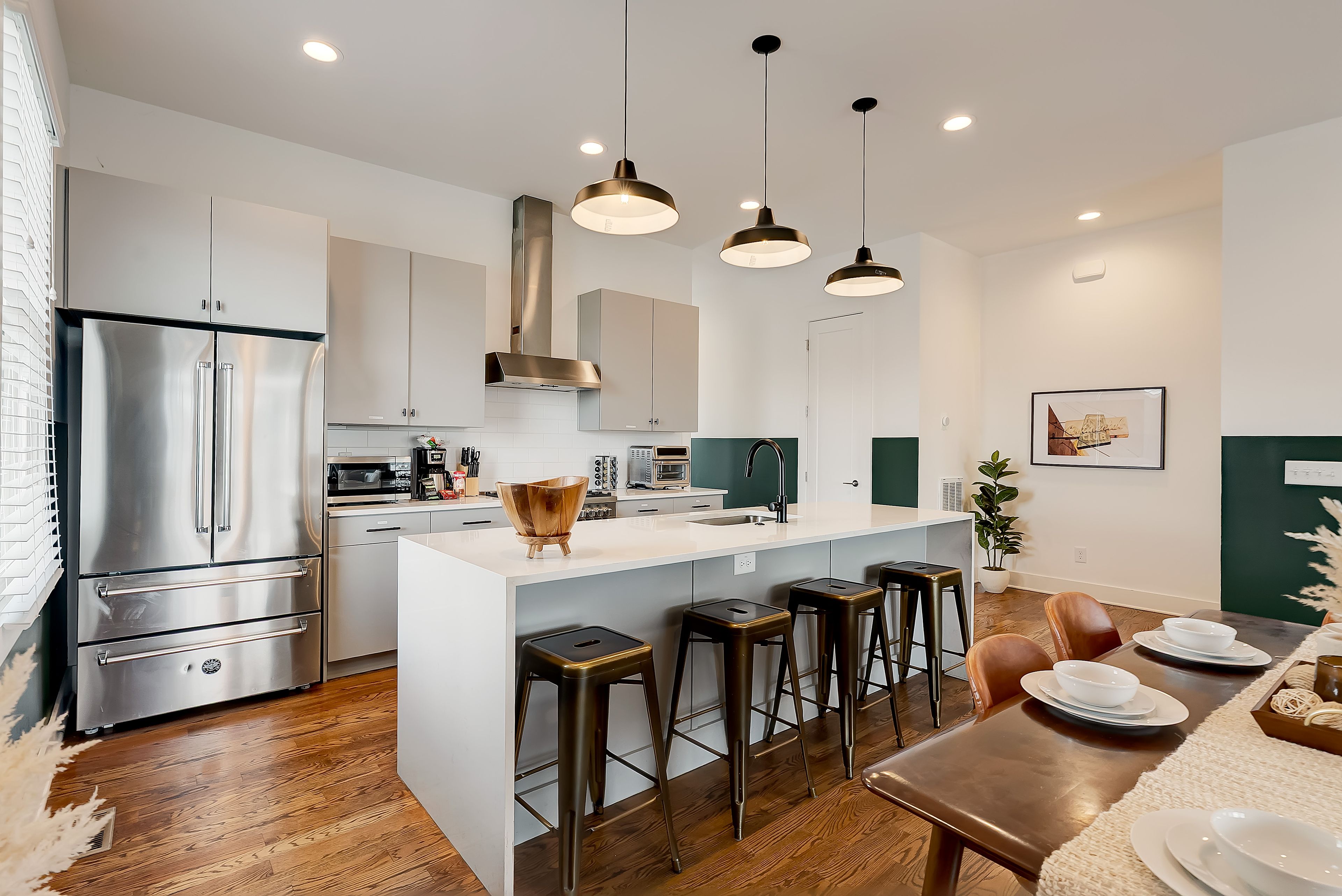 A modern kitchen with stainless steel appliances, a central island with stools, pendant lighting, and a dining area with a table set for two.