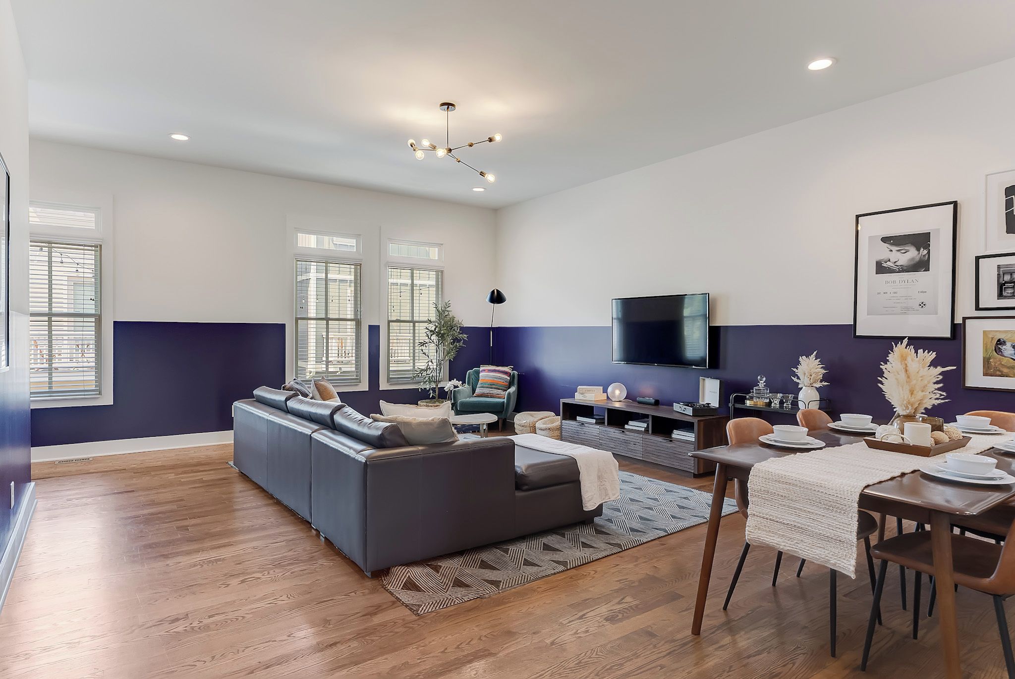 A modern living room with a grey sofa, a dining table set for four, and a flat-screen TV mounted on the wall.