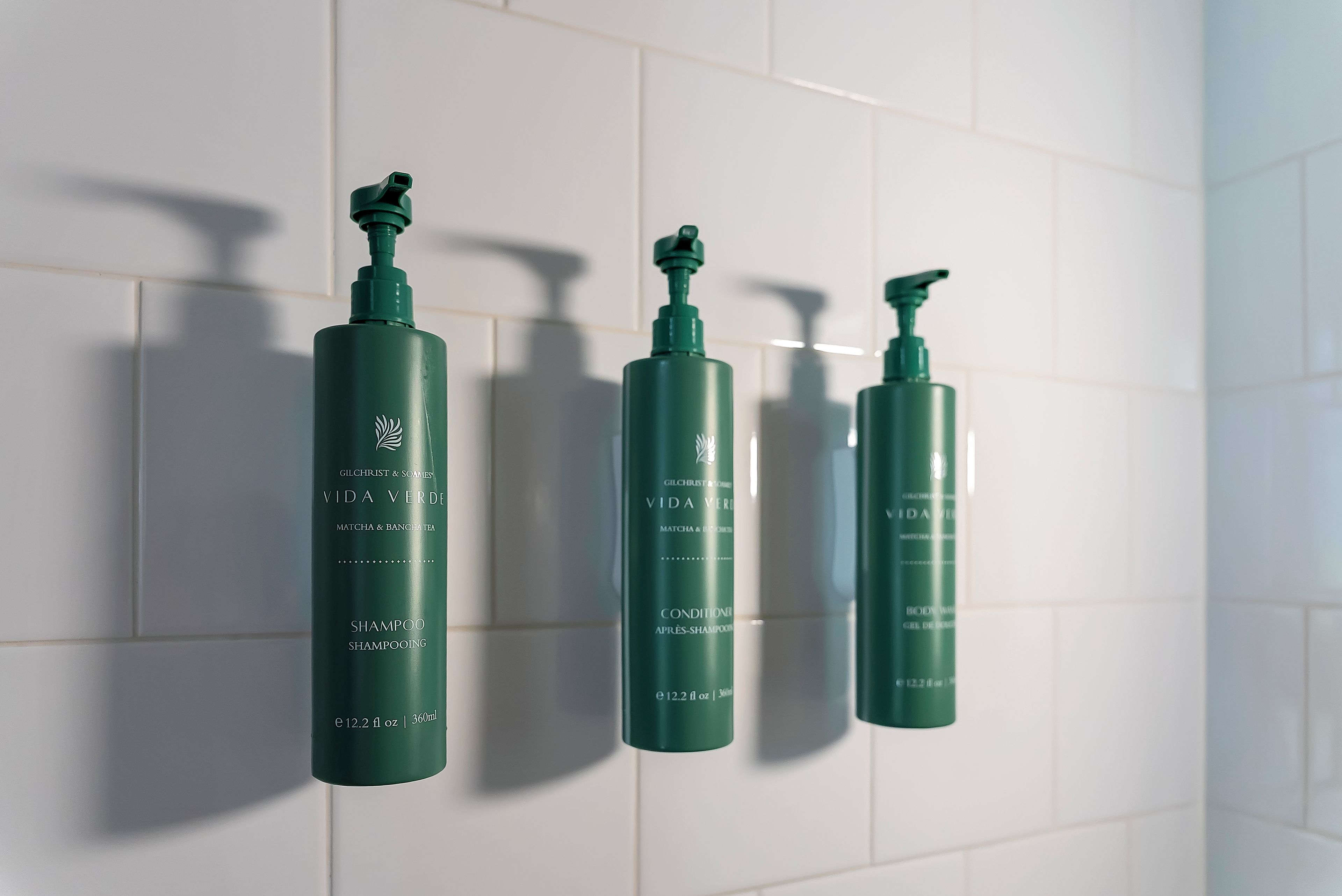 Trois bouteilles vertes de shampoing Vida Verde de la marque Gilchrist & Soames, posées sur un mur recouvert de carreaux blancs.