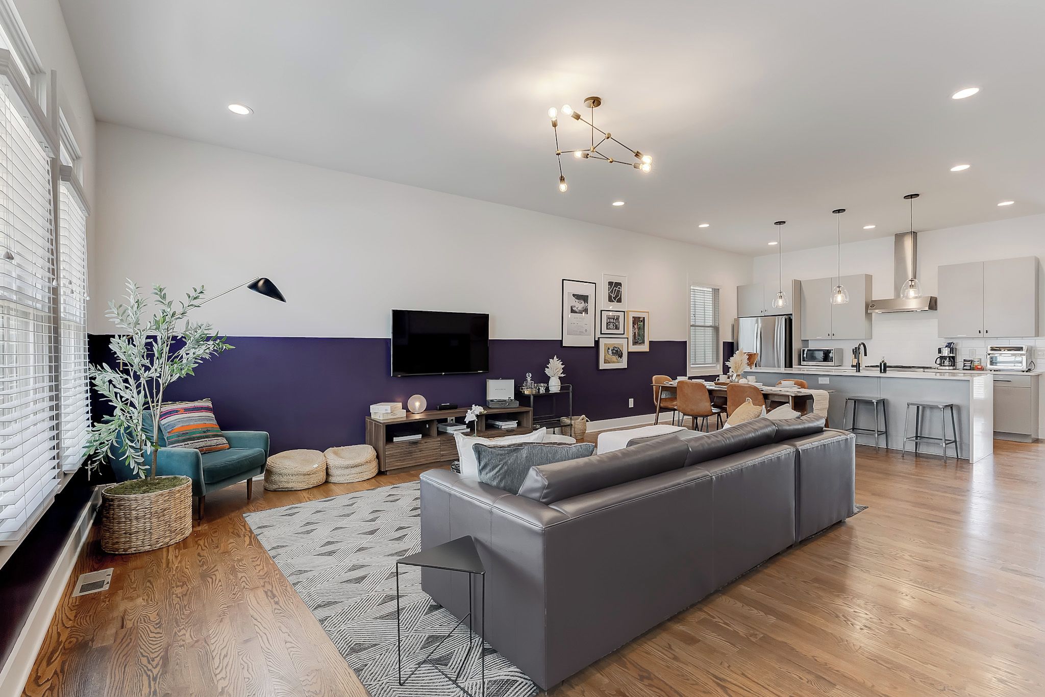 Een moderne woonkamer met een grijze bank, een televisie en een eethoek, met een keuken op de achtergrond.