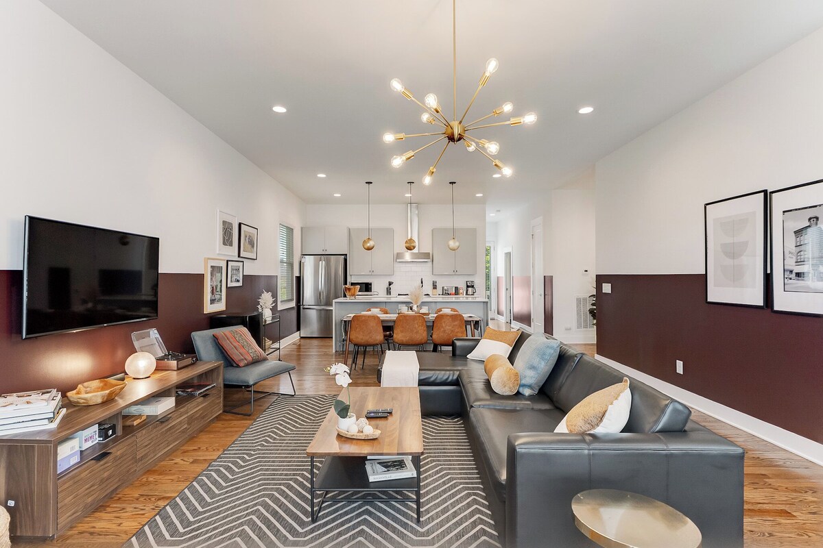 A modern living room with a sofa, coffee table, and TV. There is a kitchen in the background with a dining area.