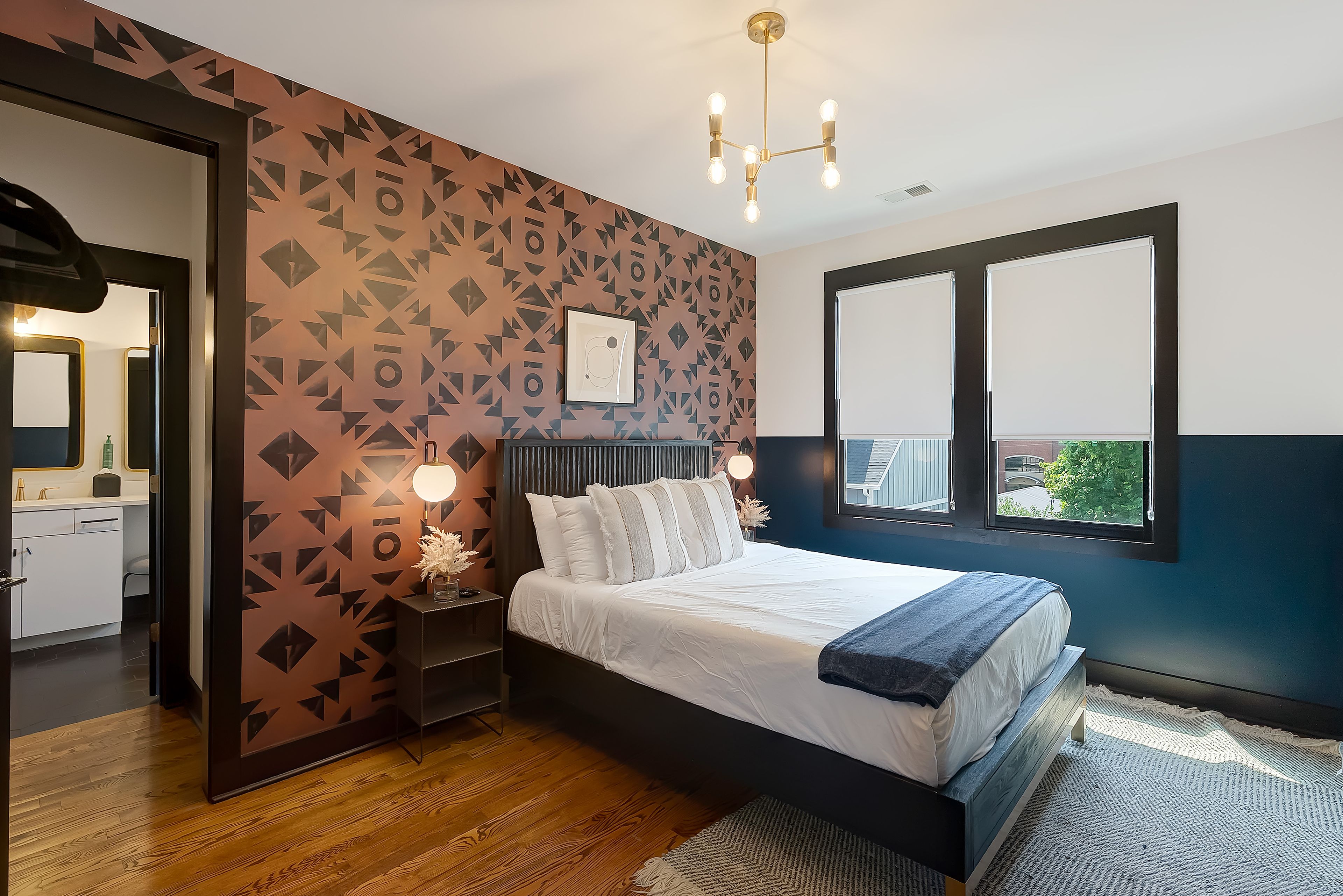 Une chambre moderne avec un mur orné d’un motif géométrique, un grand lit avec une literie blanche, une table de chevet avec une lampe, et une vue par la fenêtre.