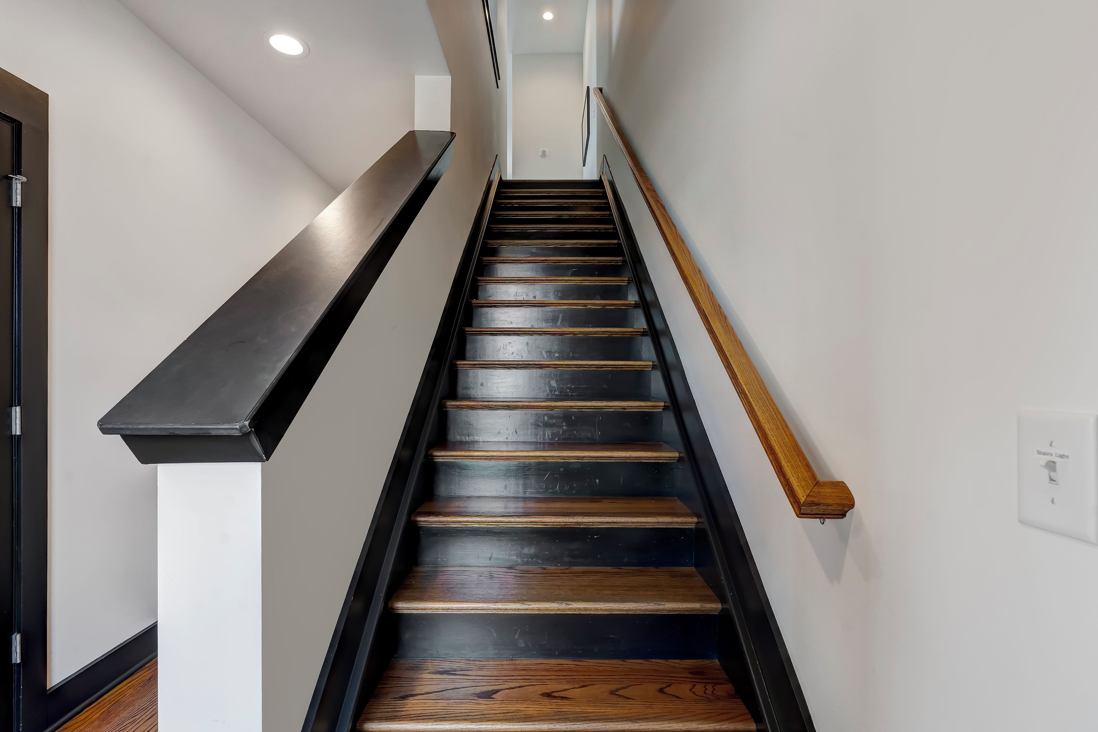 Una escalera con peldaños de madera y barandillas negras, que conduce a un rellano con una pared blanca y una puerta.