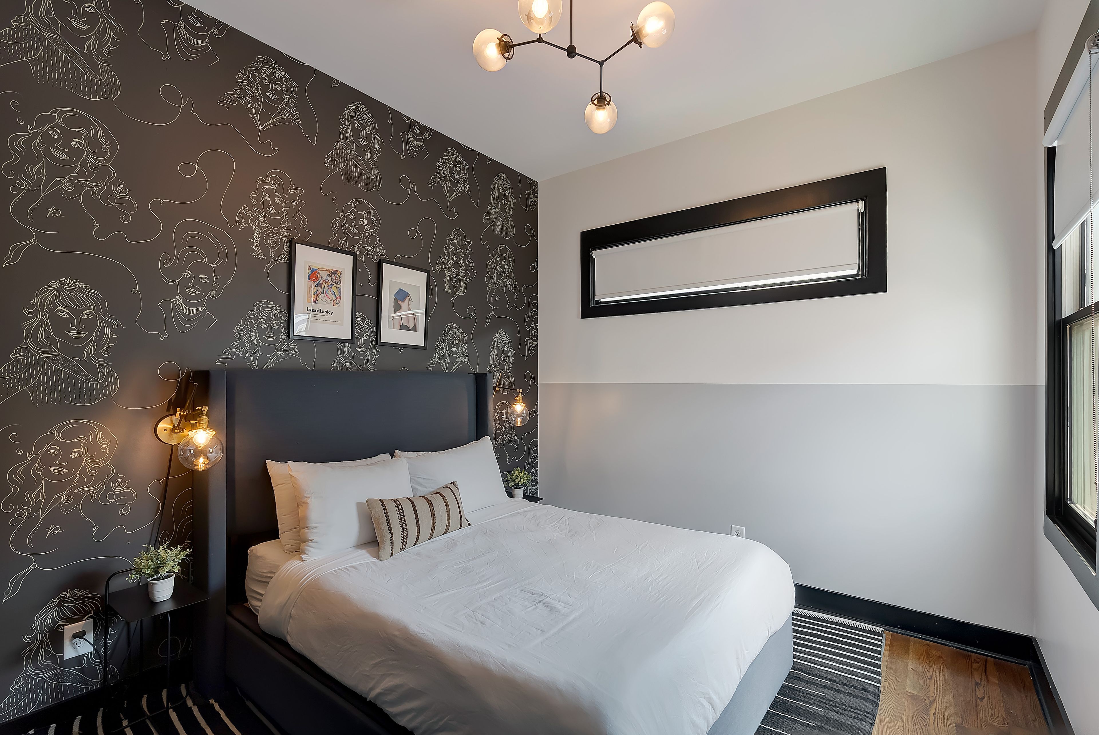 Une chambre moderne avec un grand lit, une tête de lit sombre, des œuvres d’art accrochées au mur et un luminaire fixé au plafond.