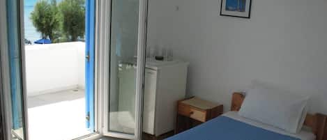 Una habitación con dos camas, un escritorio pequeño, un refrigerador y un ventanal corredero que da a un balcón con vista al mar.