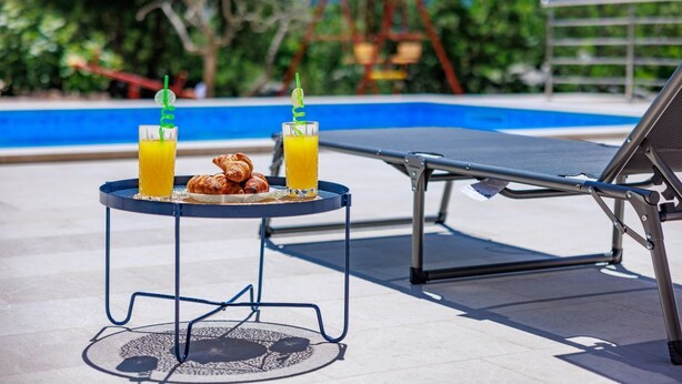 A poolside area with a lounge chair, a small table with croissants and orange juice, and a swing set in the background. - Perfect 3-bedroom house with heated pool in peaceful Šestanovac (Šestanovac)