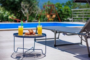 A poolside area with a lounge chair, a small table with croissants and orange juice, and a swing set in the background. - Perfect 3-bedroom house with heated pool in peaceful Šestanovac (Šestanovac)
