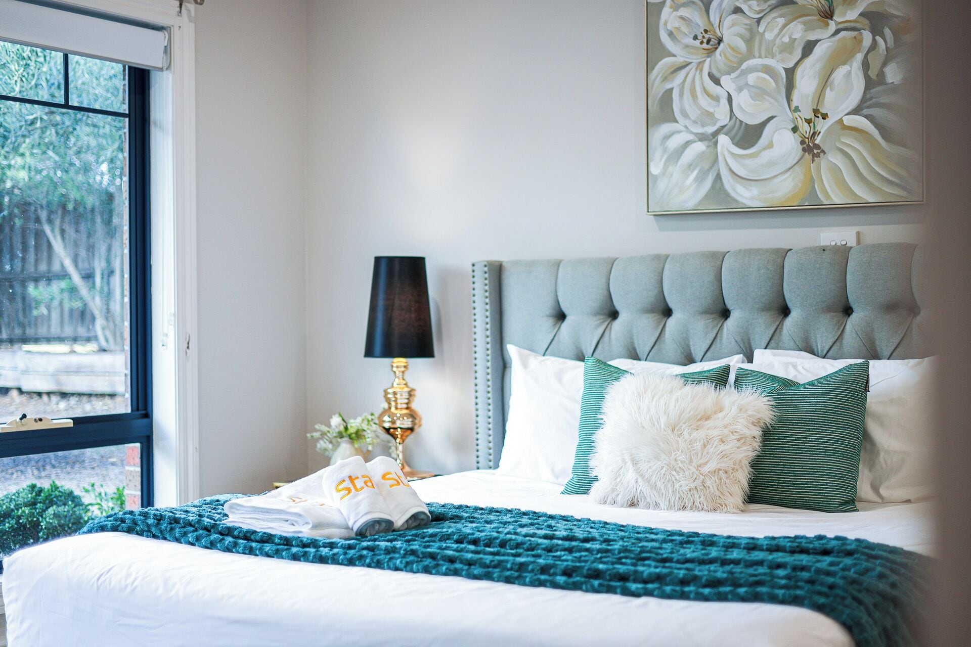 A neatly made bed with a teal blanket, white pillows, and a fluffy white pillow. There is a lamp with a black shade, a framed floral artwork on the wall, and a window with a view of greenery.