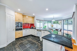 A modern kitchen with a black tiled floor, white and wooden cabinets, a built-in oven, and a large window overlooking a backyard. - StayAU PoolTable 2-Story Pet-friendly  (Caroline Springs)