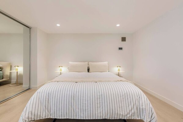 Une chambre avec un grand lit, deux lampes de chevet, un miroir et une petite grille d’aération sur le mur.