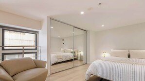 A modern bedroom with a bed, a sofa, a large mirror, and a window. - Luxuria Apartments - Degraves Place (Melbourne)