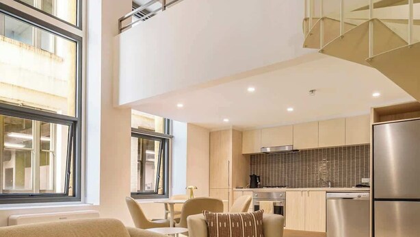 A modern living room with a sofa, ottoman, and a dining area with chairs. - Luxuria Apartments - Degraves Place (Melbourne)