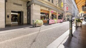 A street with a building featuring a pizza bar, a store with a pink sign, and a potted plant on the sidewalk. - Luxuria Apartments - Degraves Place (Melbourne)