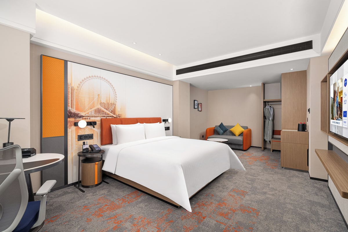 A hotel room with a bed, a desk, a chair, a small table, a trash bin, a sofa, a TV, and a coat rack.