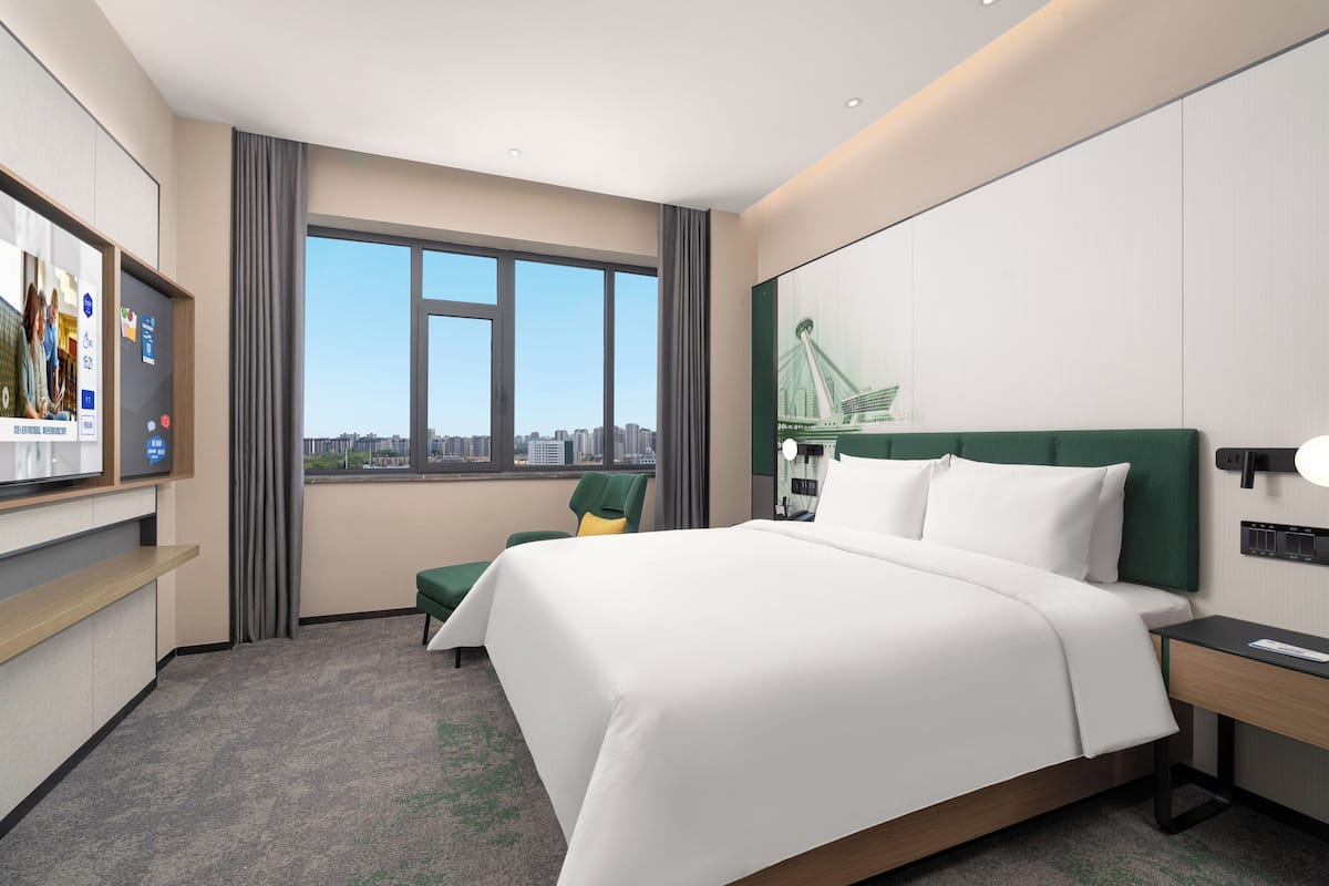 A hotel room with a large bed, a desk, a chair, a TV, and a window with a city view.