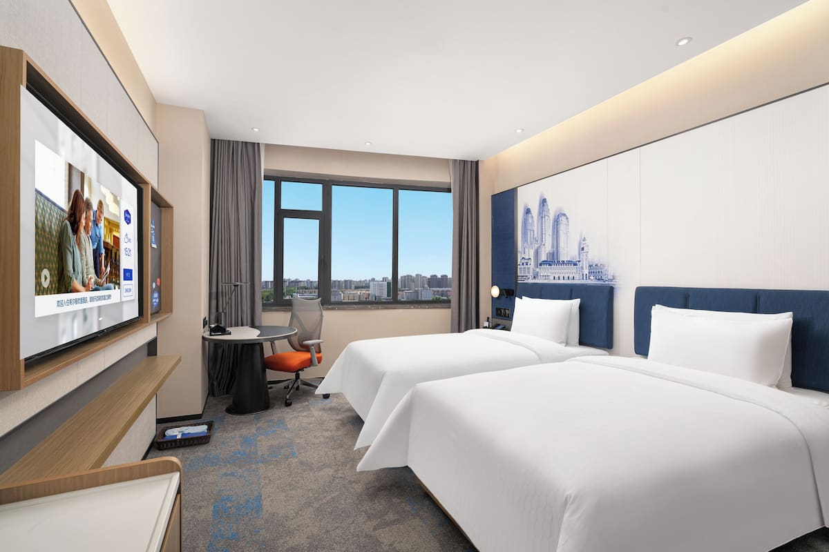 A hotel room with two beds, a desk with a chair, a television, and a large window with a city view.