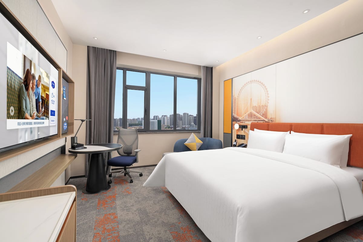 A hotel room with a bed, a desk, a chair, a TV, a window with a city view, and a large poster on the wall.
