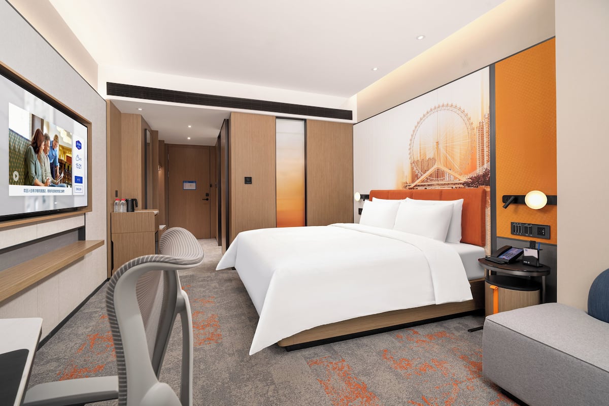 A hotel room with a bed, a desk, a chair, a TV, and a large window with a view of a Ferris wheel.