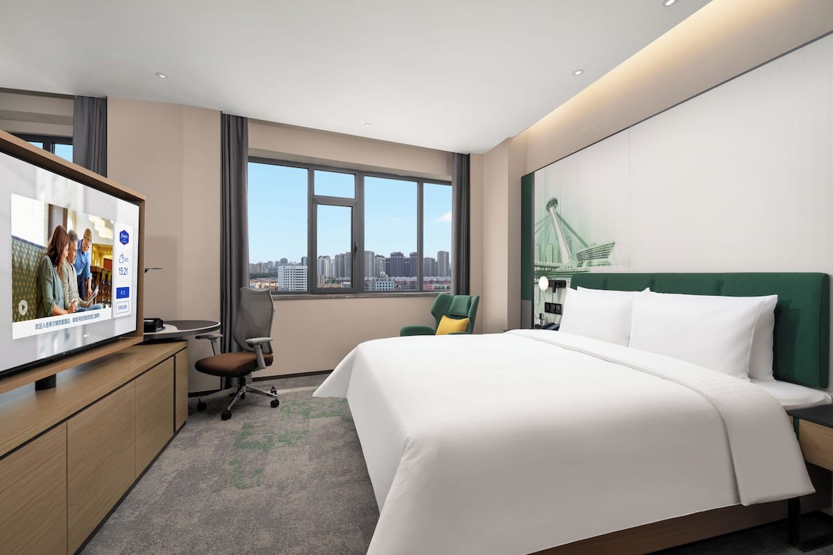 A hotel room with a bed, a desk, a chair, a TV, and a window with a city view.