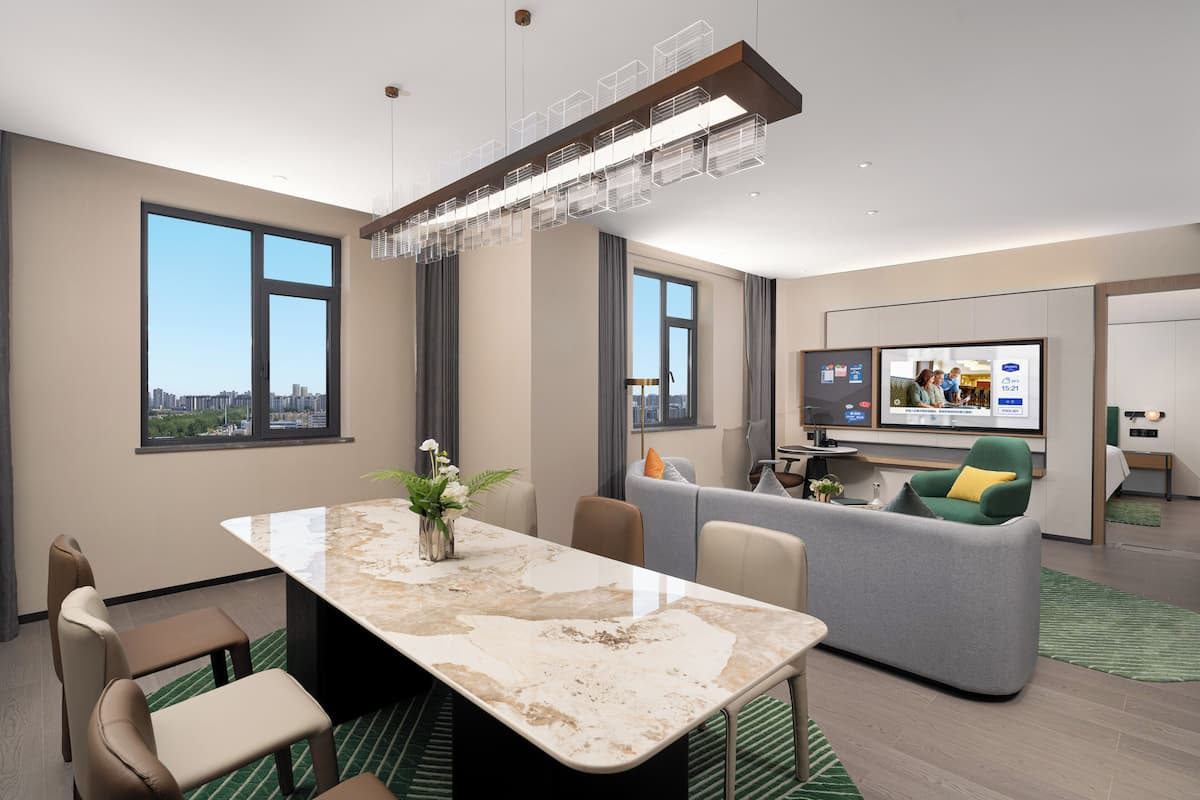 A modern hotel room with a marble dining table, a sofa, a TV, and large windows.