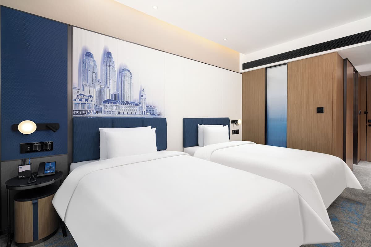 A hotel room with two beds, a cityscape artwork on the wall, a desk with a phone, and a trash bin.