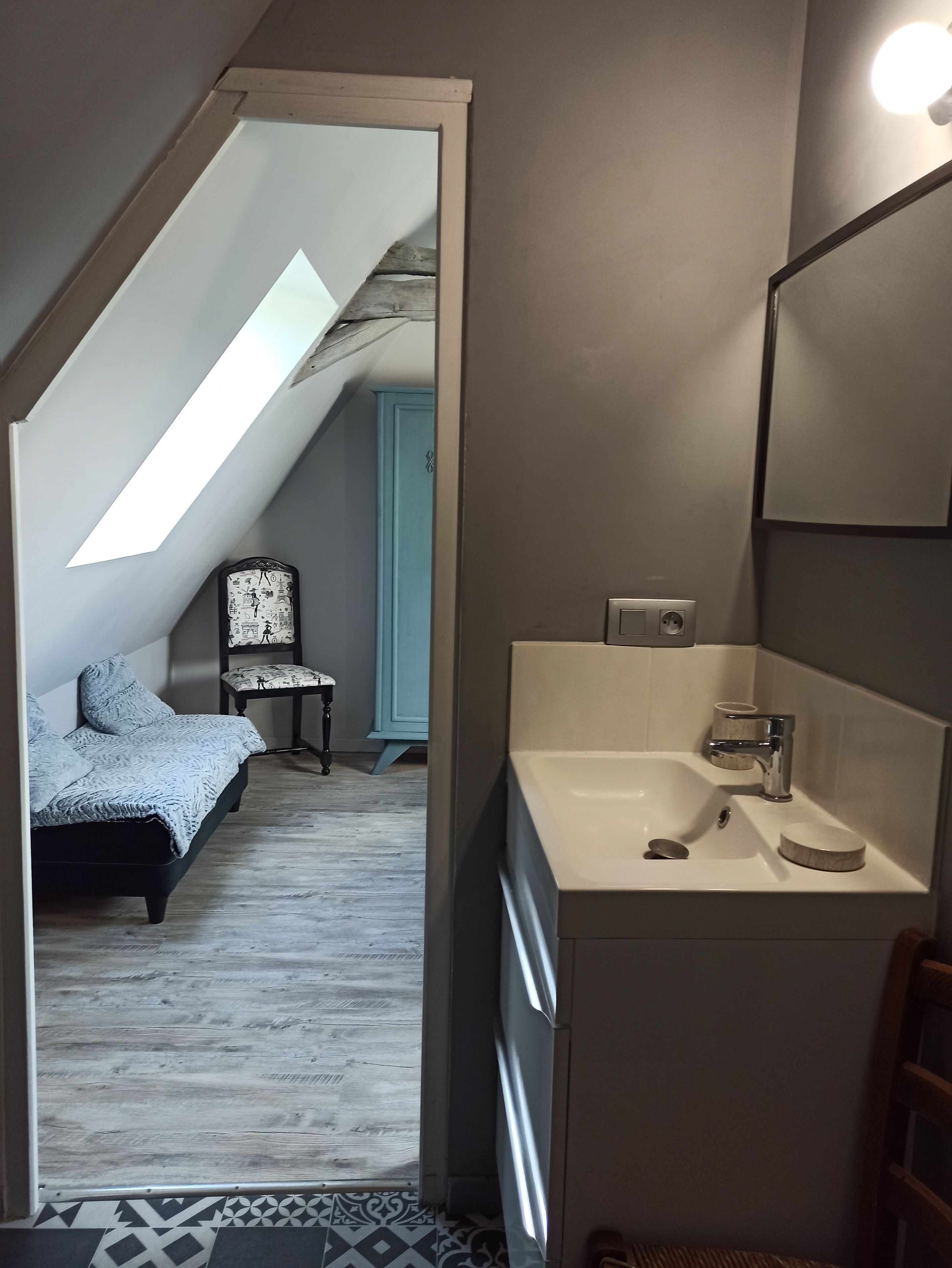 Een slaapkamer met schuine wand, een bed, een stoel, en een badkamer met wastafel en spiegel.
