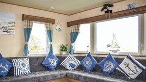 Ein gemütliches Wohnzimmer mit grauem Sofa, verziert mit nautisch inspirierten Kissen, einer Holzvertäfelung und einem Fenster mit Blick auf ein Segelboot.