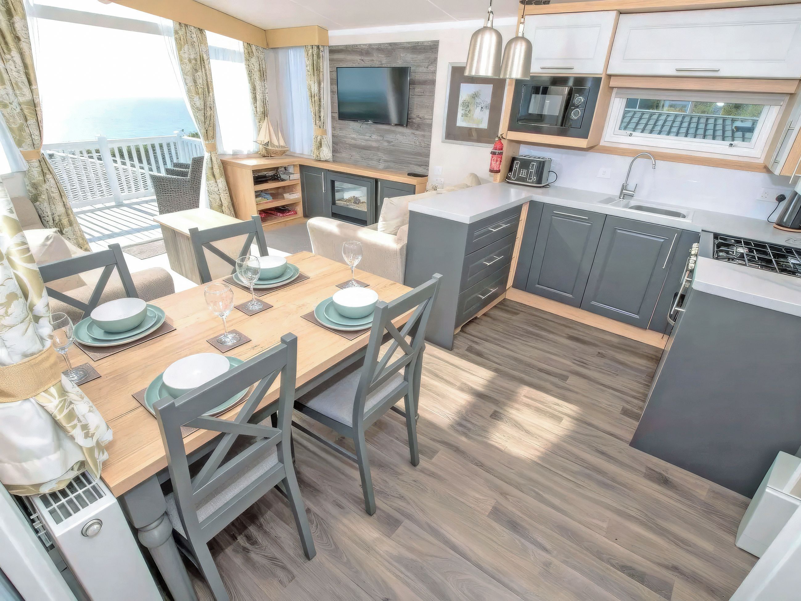 A modern kitchen with grey cabinets, a wooden dining table set for four, and a view of the ocean through large windows.