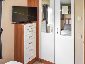 Ein Schlafzimmer mit Fernseher auf einer Holzkommode, einem Kleiderschrank mit Spiegel und einem Bett mit rosa-weißer Karodecke.