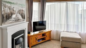 Ein Wohnzimmer mit Kamin, einem Fernseher auf einem Holzboden und einem beigefarbenen Sofa, sowie einem großen Fenster mit transparenten Vorhängen.