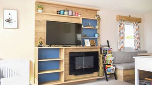A living room with a TV, fireplace, shelves with items, and a sofa. - Juniper Close 52 - UKC9405 (Exmouth)
