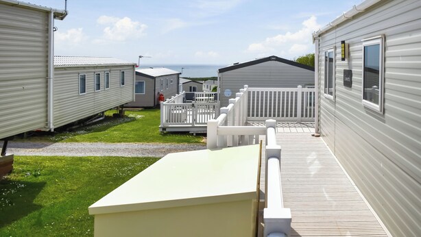 A row of mobile homes with a grassy area in front, a white fence, and a clear sky. - Juniper Close 52 - UKC9405 (Exmouth)