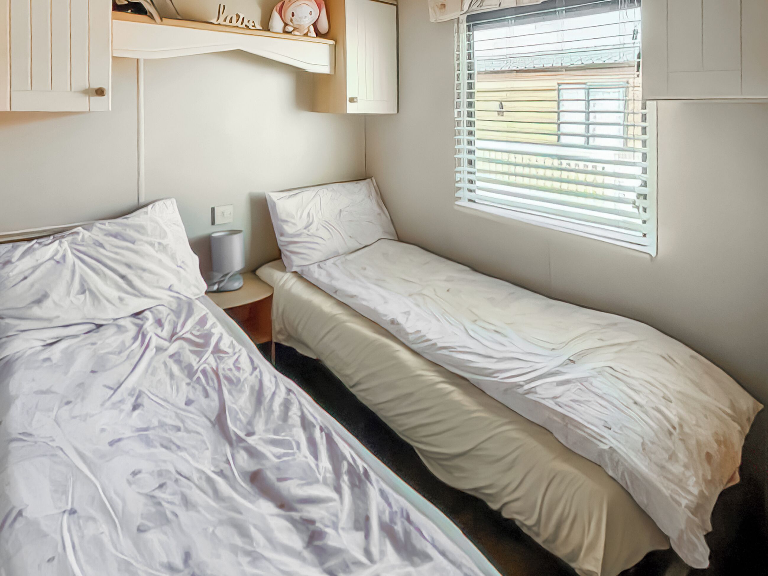 A room with two single beds, a small bedside table, a shelf with decorative items, and a window with blinds.