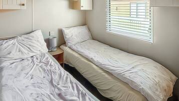 A room with two single beds, a small bedside table, a shelf with decorative items, and a window with blinds.