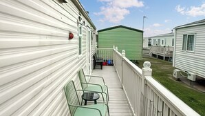 A covered outdoor area with a wooden deck, metal chairs, and a green building in the background.