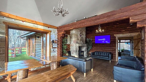 A rustic cabin interior with a stone fireplace, wooden benches, a black leather sofa, and a television screen displaying 'Smooth.' - Bellstane Chalet - UKC9440 (Glasgow)