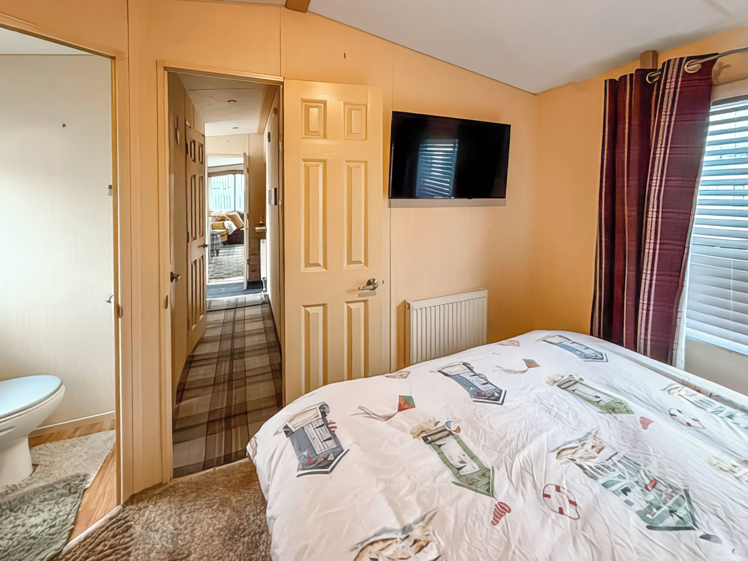 Un dormitorio con cama, televisor montado en la pared, puerta que da a otra habitación y radiador.