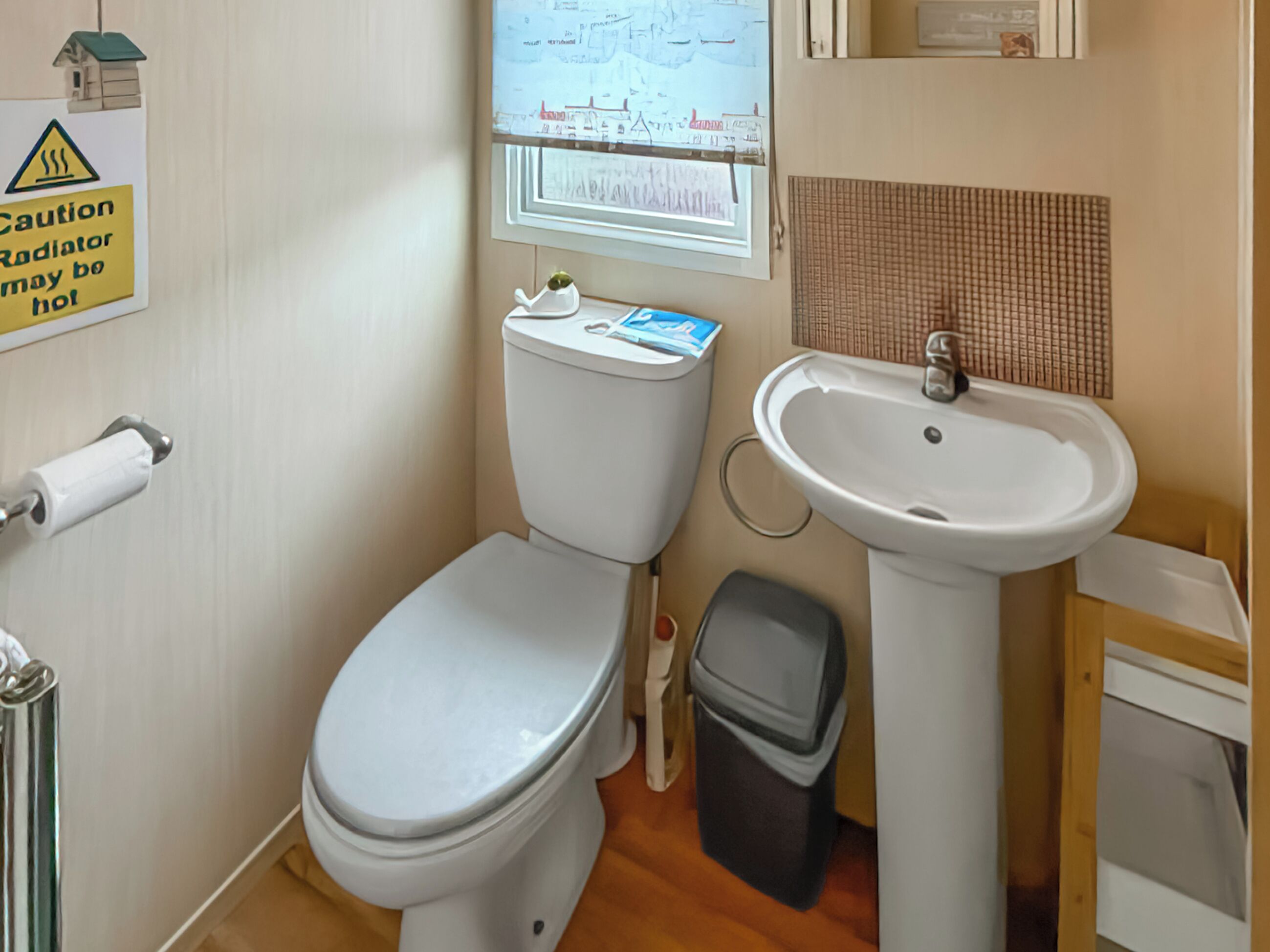 A bathroom with a toilet, sink, and trash bin. There is a caution sign on the wall.