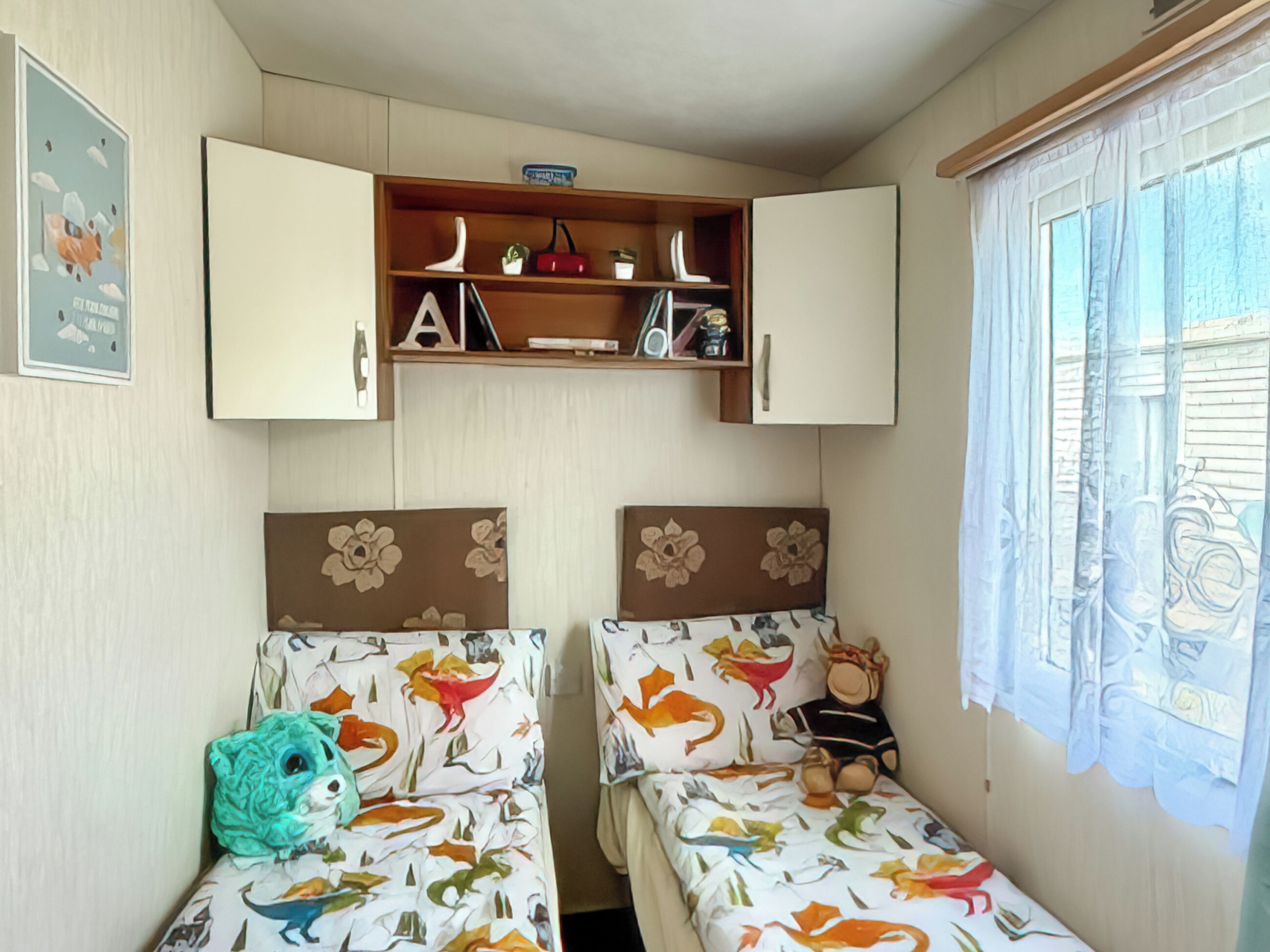 A room with two beds, a shelf with books and decorative items, and a window with curtains.