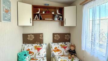 A room with two beds, a shelf with books and decorative items, and a window with curtains.